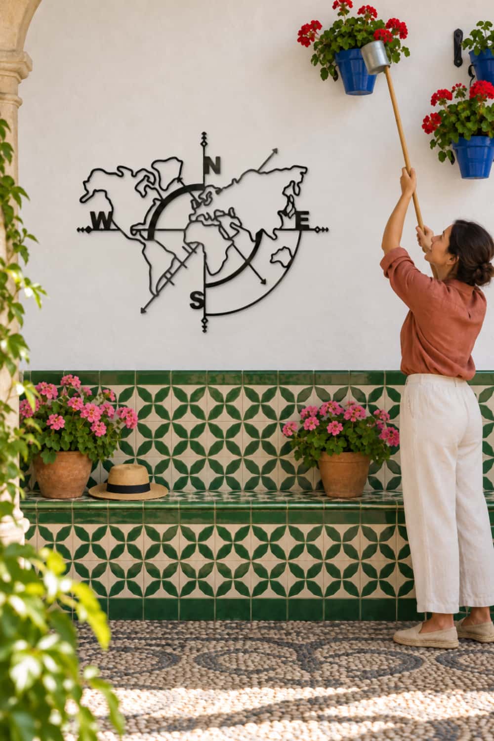 Mapa Mundi de aluminio negro en un patio de Córdoba mientras una mujer riega las macetas de geranios